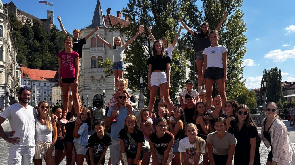 Skupinska fotografija udeležencev tabora na Prešernovem trgu v Ljubljani, ki izvajajo cheerleading dvige; v ozadju se vidita Tromostovje in Ljubljanski grad.